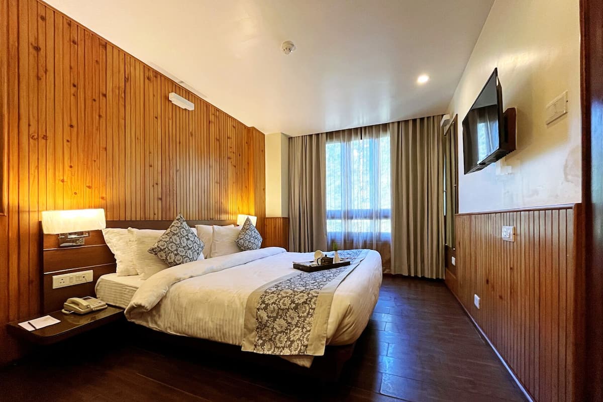 A hotel room with a wooden paneled wall, a bed with a patterned bedspread, a bedside table with a lamp, a flat-screen TV mounted on the wall, and a window with curtains.