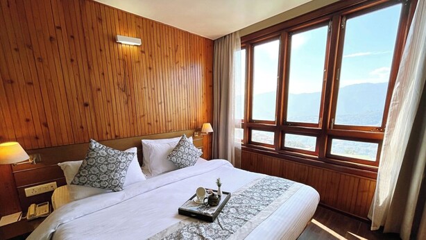 A hotel room with a large bed, wooden panel walls, a nightstand with a lamp, and a window with a view of the sea. - Cilantro Unwind Delisso Abode (Gangtok)