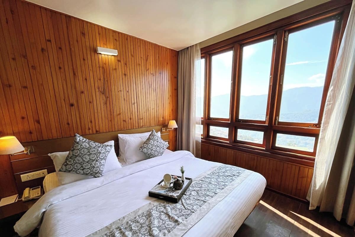 A hotel room with a large bed, wooden panel walls, a nightstand with a lamp, and a window with a view of the sea.