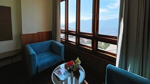 A hotel room with a large window offering a mountain view, a blue armchair, a small round table with a magazine and flowers, and a wooden headboard. - Cilantro Unwind Delisso Abode (Gangtok)