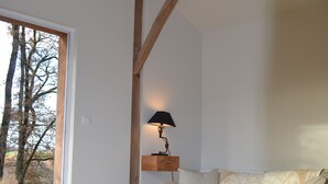 A living room with a wooden ceiling, a white sofa with patterned cushions, a small table with a candle and a vase, and a view of trees outside. - Gîte 4 persons Mathys (Doissat)