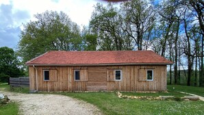 A wooden building with a red roof, a gravel driveway, and a grassy area with trees in the background. - Gîte 4 persons Mathys (Doissat)