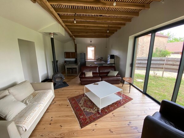 A modern living room with a sofa, a coffee table, and a fireplace. There is a kitchen area with cabinets and a refrigerator. The room has large windows and a wooden ceiling.