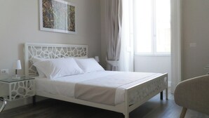 A neatly made bed with white linens in a room with a modern design, including a geometric headboard and a glass side table with a lamp.
