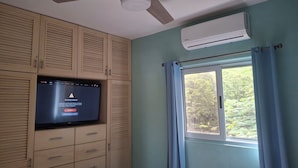 Una habitación con ventilador de techo, aire acondicionado, televisor montado en un mueble de madera y una ventana con cortinas.