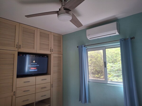 A room with a ceiling fan, an air conditioning unit, a television mounted on a wooden cabinet, and a window with curtains.