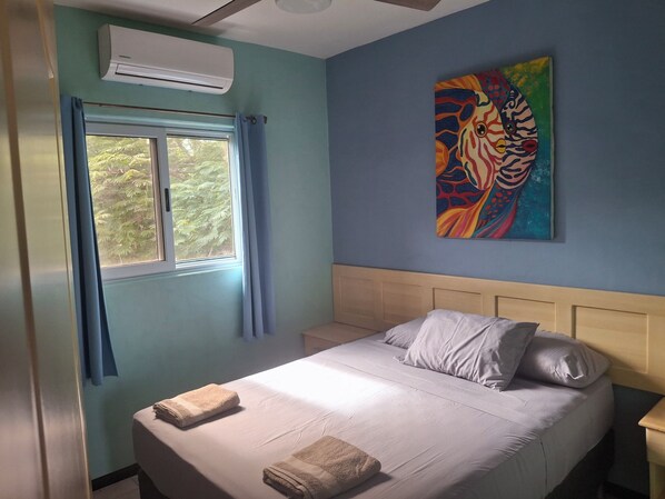 A bedroom with a bed, pillows, a painting, an air conditioner, and a window with curtains.