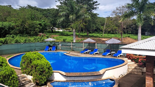Una piscina con agua azul, rodeada de arbustos y árboles verdes. Hay sillas de descanso azules y sombrillas a rayas.