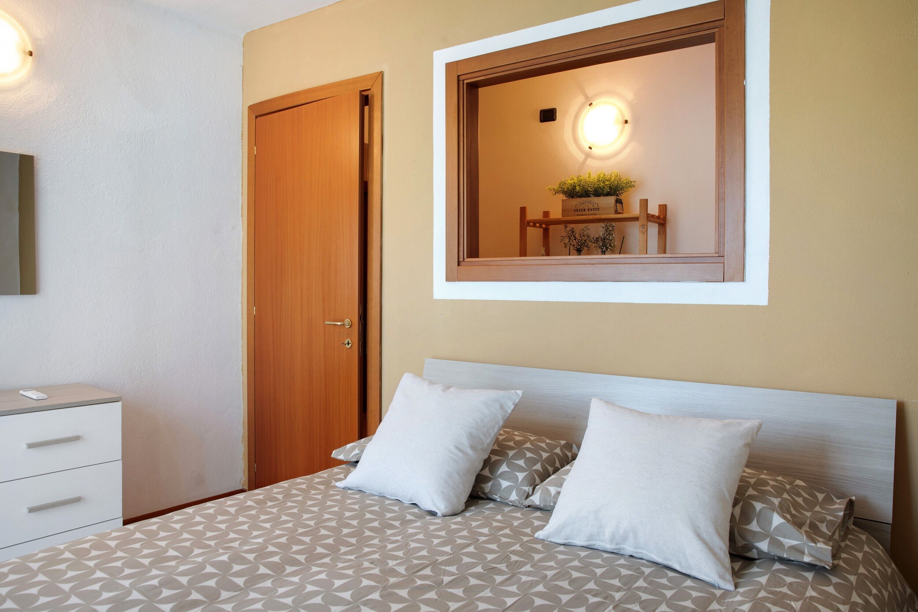 A bedroom with a bed, pillows, a nightstand, a dresser, a mirror, and a wall-mounted light fixture.