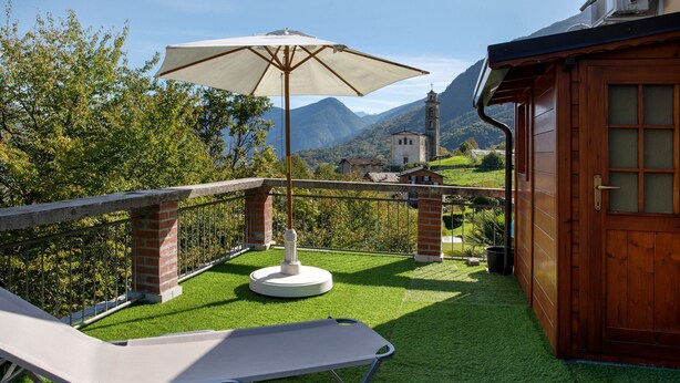 A balcony with a lounge chair, a table with an umbrella, and a view of mountains and a church. - Apartment 'La Terrazza' with Mountain View, Wi-Fi and Air Conditioning (Nesina)