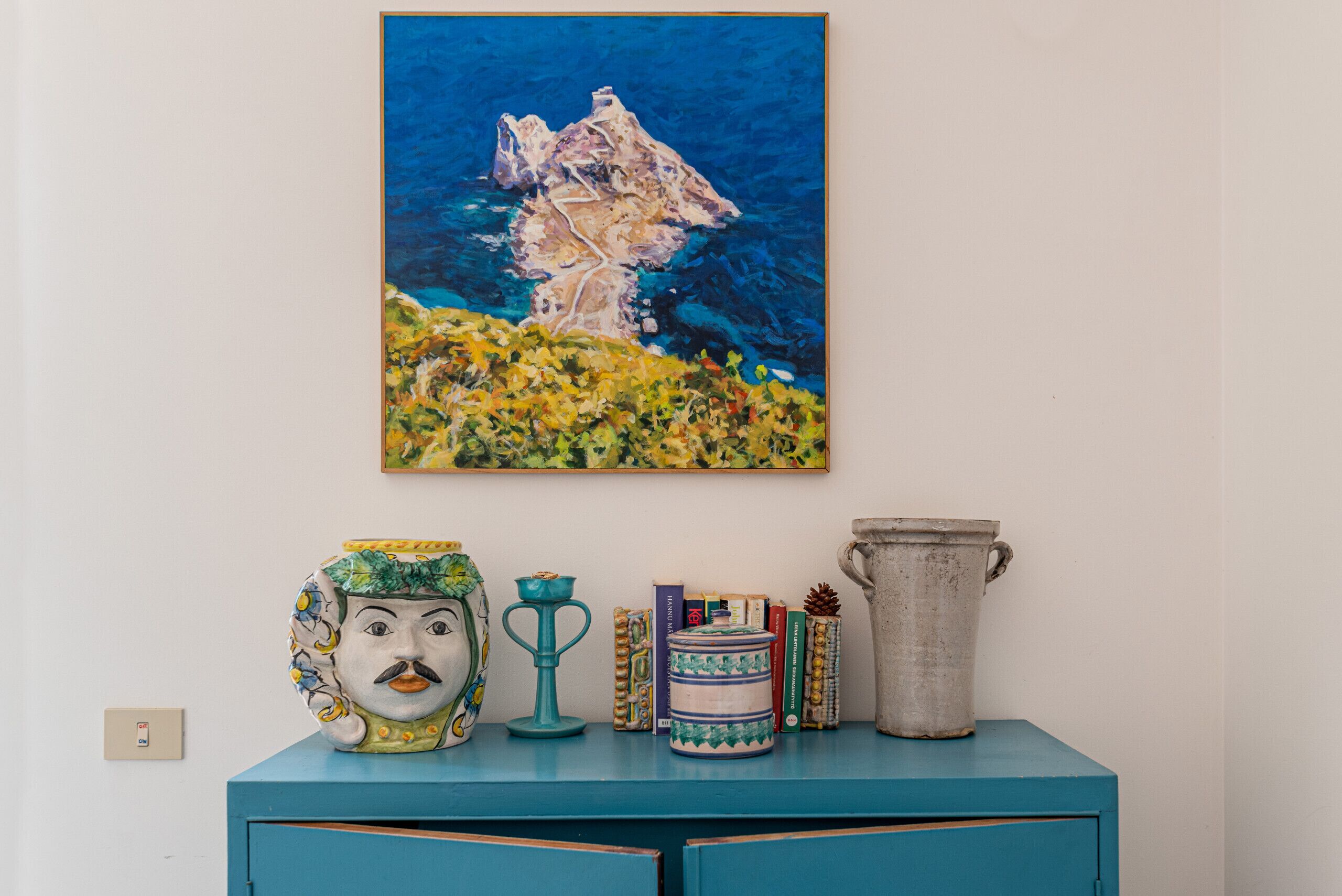 Un mobile color acquamarina con un dipinto di un'isola rocciosa, una brocca in ceramica, un vaso decorativo e diversi libri.