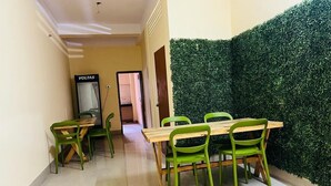 A dining area with a wooden table and green chairs, a green hedge wall, and a sign reading "VOLTAS". - Hotel Cosy Home (Guwahati)