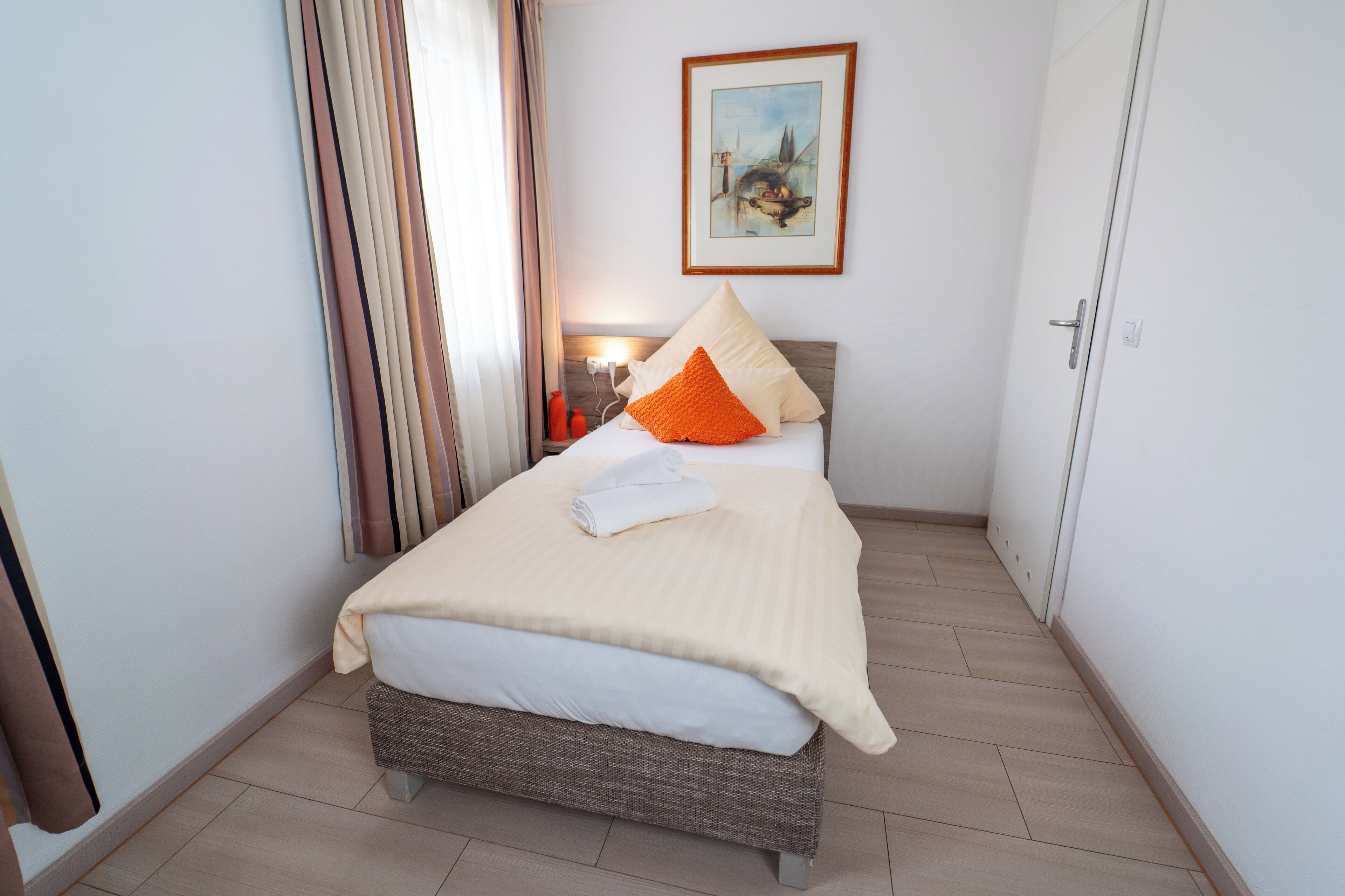 A neatly made bed with a white and orange pillow, a beige blanket, and a folded towel. There is a framed picture on the wall above the bed.