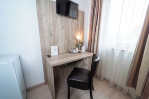 Quarto pequeno e moderno com uma escrivaninha de madeira, uma cadeira preta, televisão de tela plana na parede e uma janela com cortinas finas.