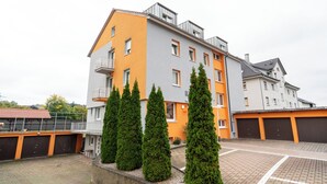 Ein mehrgeschossiges Wohngebäude mit einer Kombination aus orangefarbenen und grauen Außenwänden, einem Balkon im oberen Geschoss sowie einem Parkplatz mit Pflasterbelag.