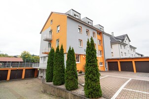 Um edifício residencial de vários andares com paredes externas em tons de laranja e cinza, uma varanda no andar superior e uma área de estacionamento com piso de tijolos.