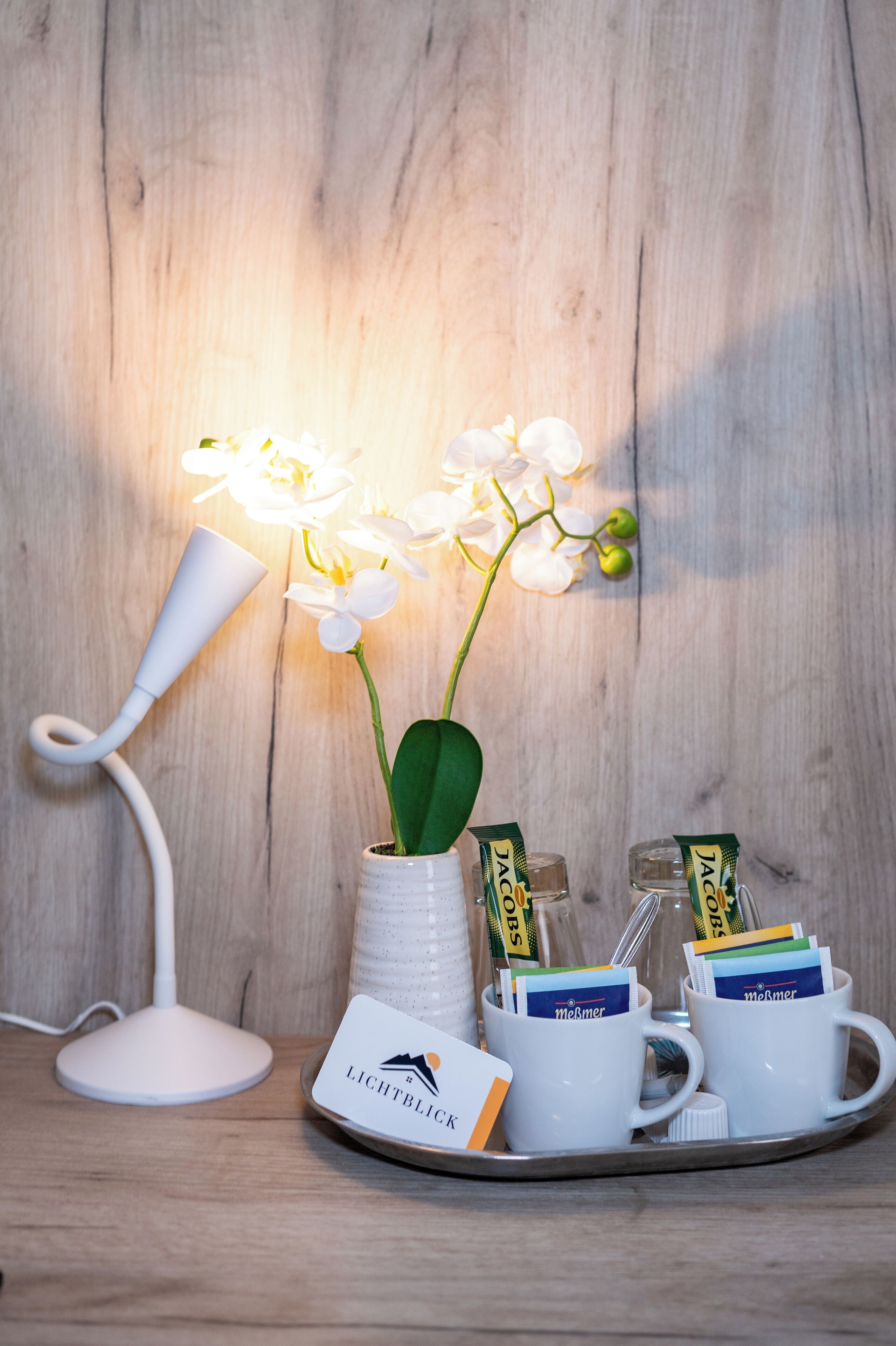 Uma bandeja com uma caneca branca, um vaso branco com uma orquídea e um abajur branco sobre uma superfície de madeira.