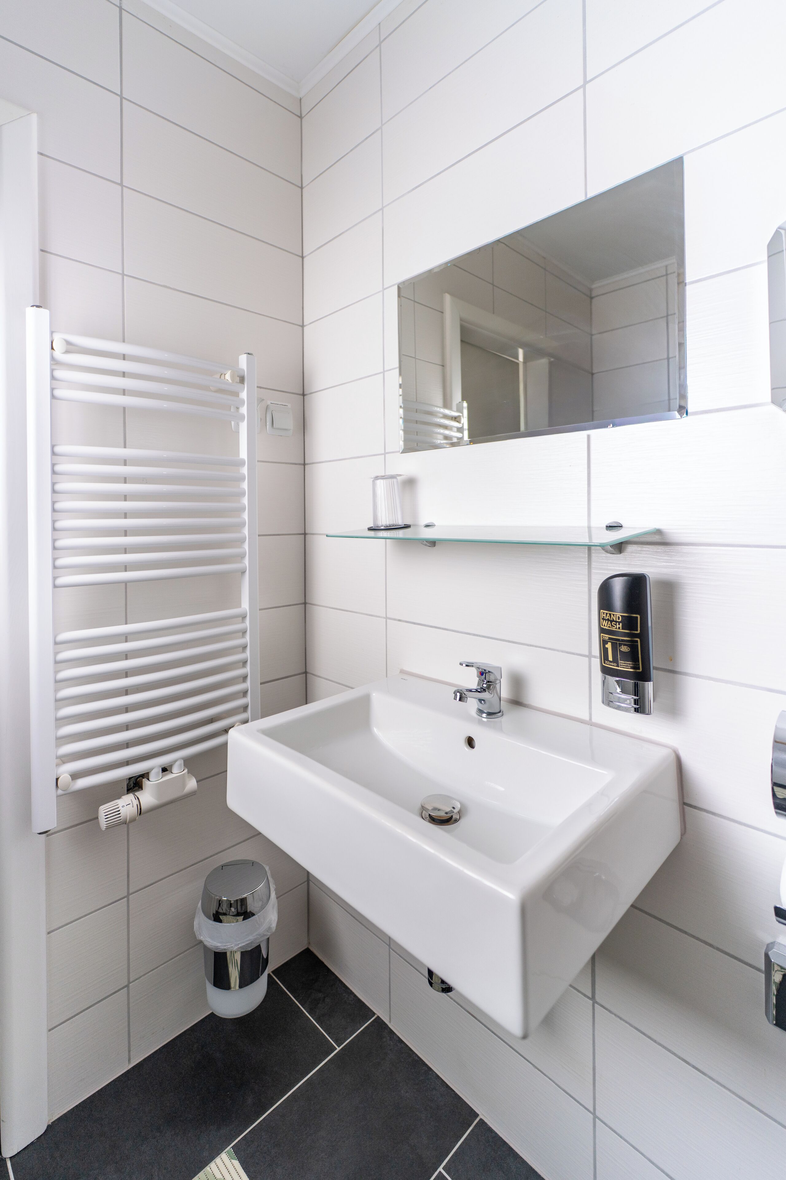 Un bany modern amb un lavabo blanc, una prestatgeria de vidre, un para-tovellons, un cendrer i un dosificador de gel hidroalcohòlic.