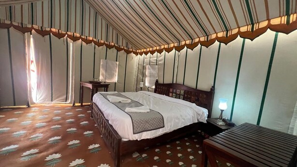 A room with a bed, a wooden nightstand, a table, and a lamp. - Shourya Desert Camp (Jaisalmer)