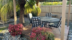Eine überdachte Terrasse im Freien mit Tisch und Stühlen, umgeben von Zierpflanzen und einem Holzzaun.