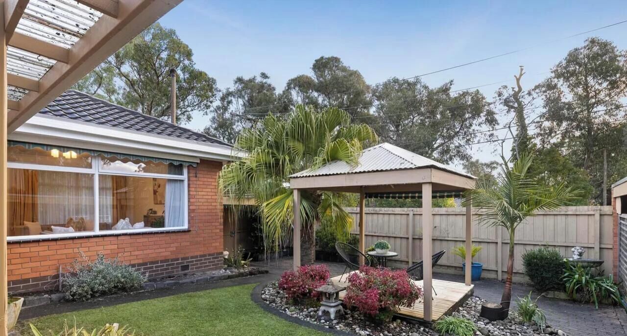 Un jardin à l’arrière d’une maison, avec une maison en briques, une pergola en bois, un kiosque et une pelouse bien entretenue agrémentée de différentes plantes.