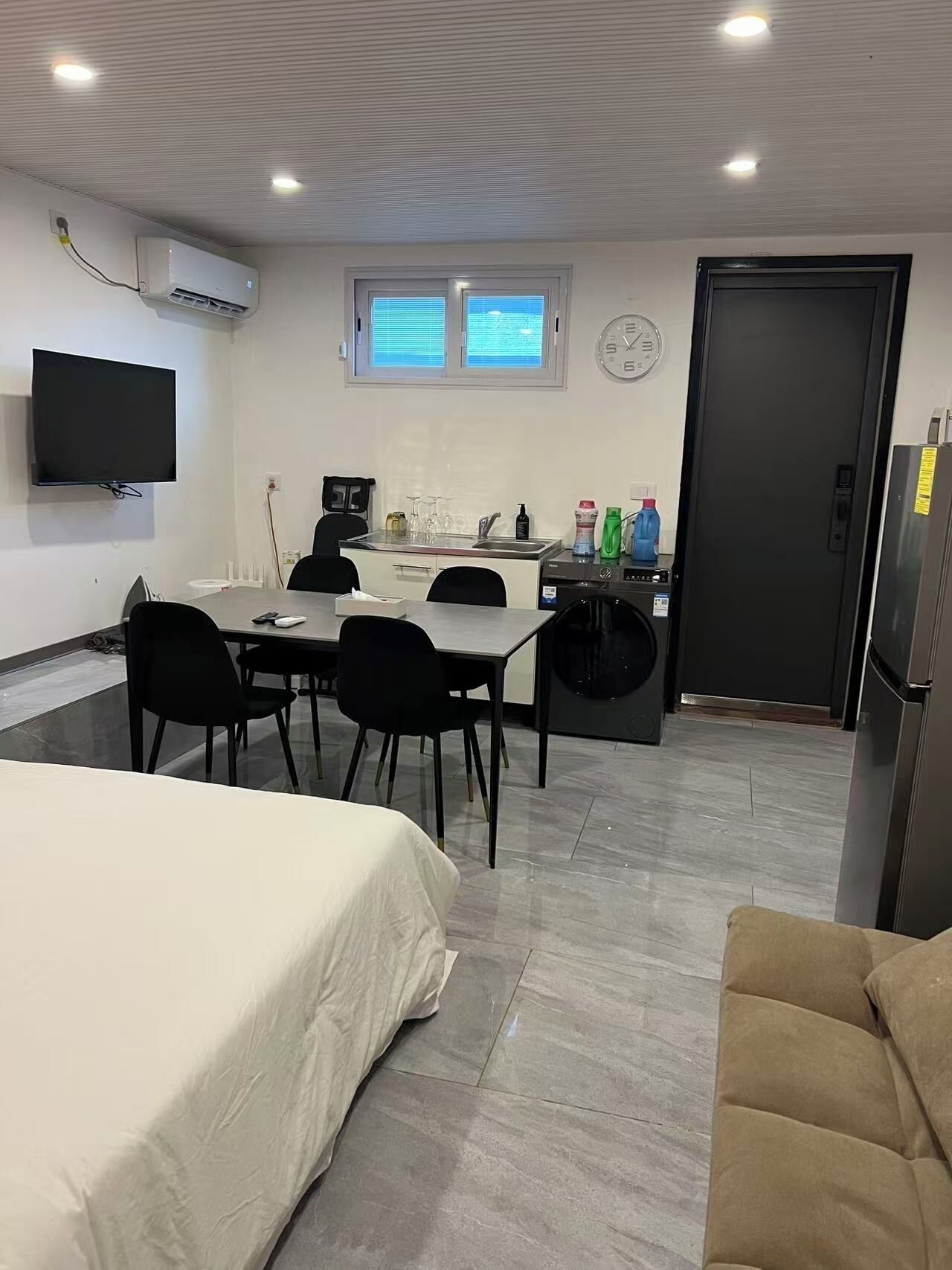 A compact hotel room with a bed, a dining table with chairs, a small kitchen area, and a TV mounted on the wall.