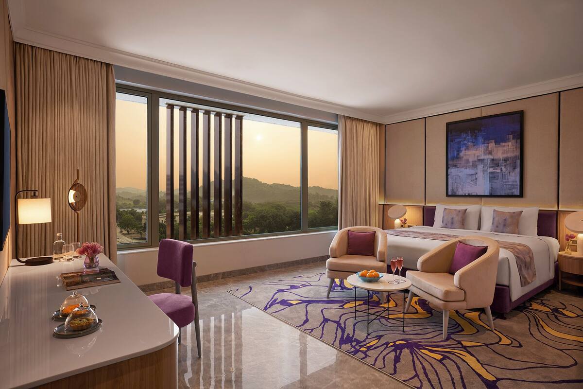 A hotel room with a large window, a bed, a desk with a chair, and a small table with a drink.