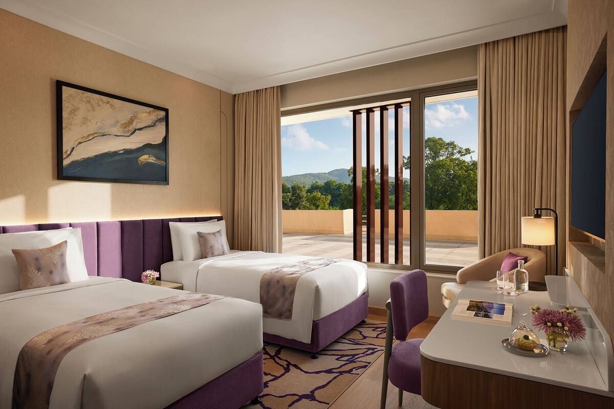 A hotel room with two beds, a desk, a chair, a lamp, and a view of the outdoors.