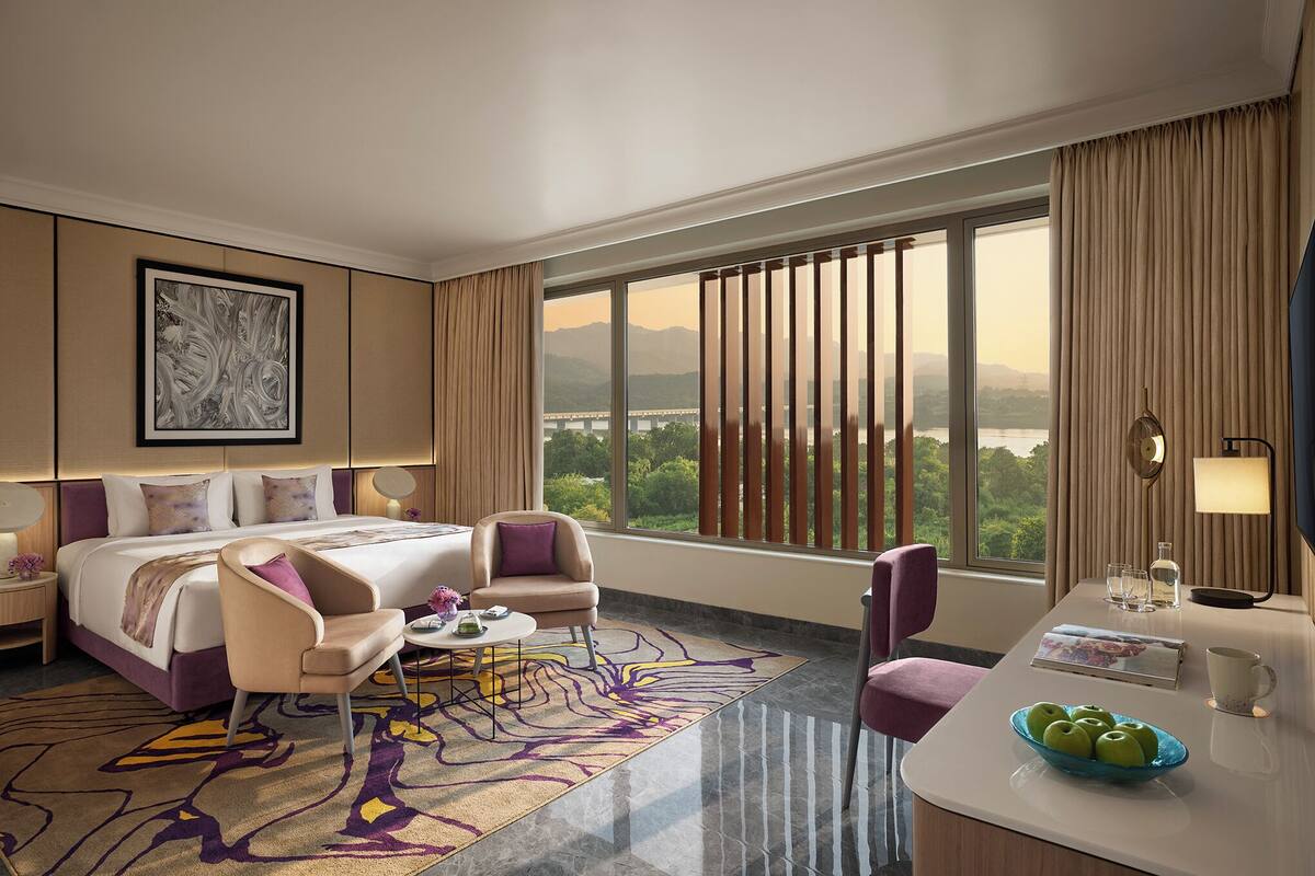A hotel room with a bed, two chairs, a small table, a coffee table, a view of mountains, and a large window with curtains.