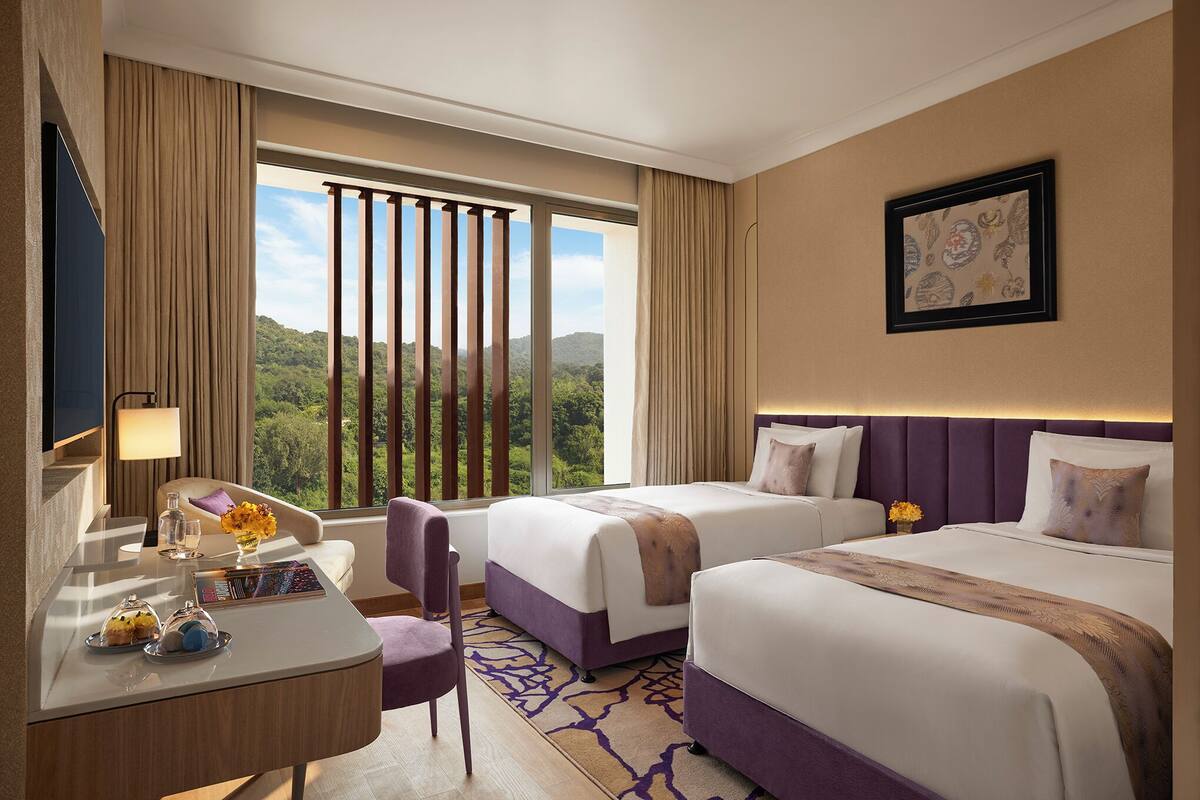 A hotel room with two beds, a desk with a lamp, a chair, and a view of the outdoors.
