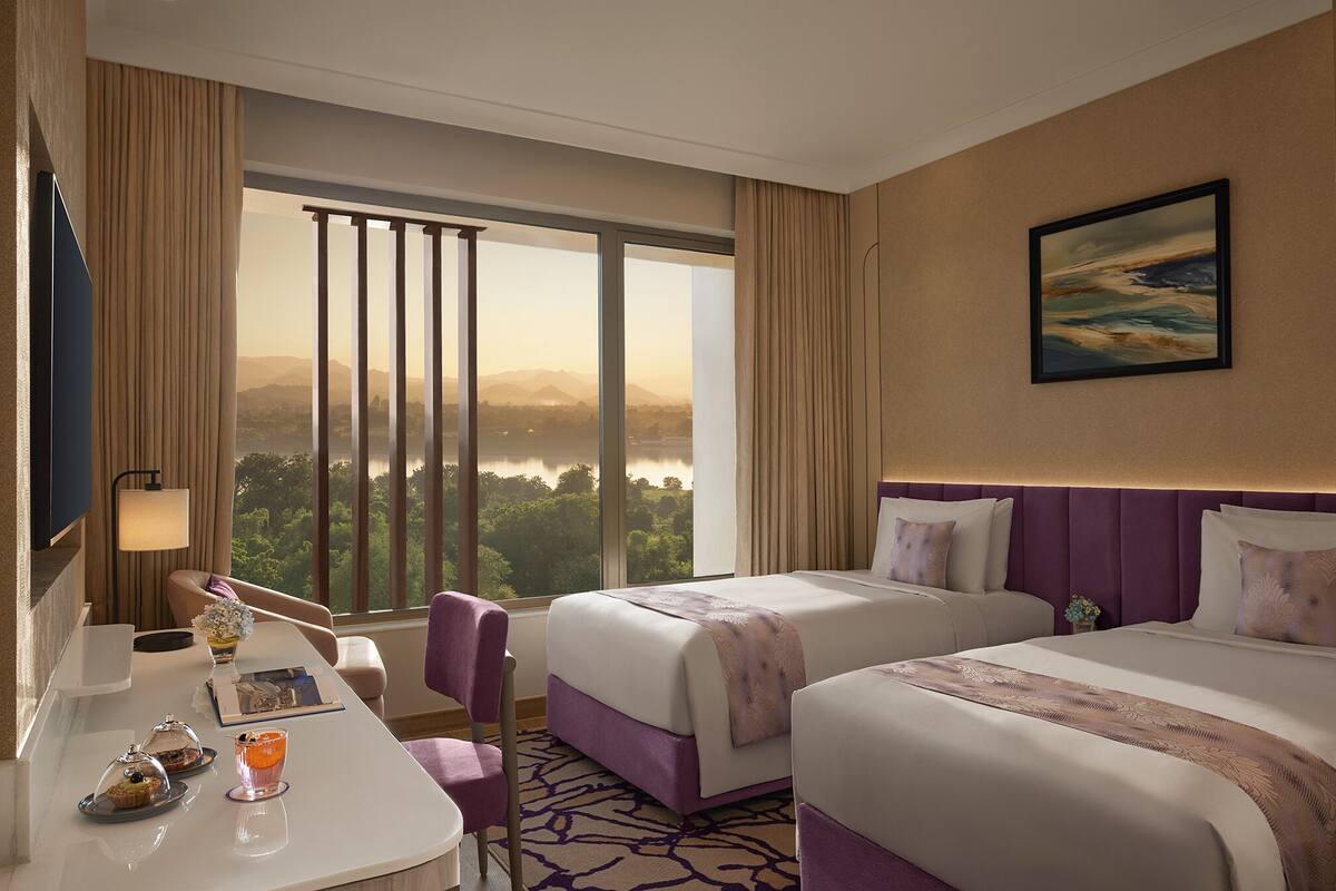 A hotel room with two beds, a desk with a chair, a view of a cityscape, and a large window with curtains.