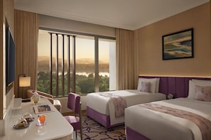 Habitación de hotel con dos camas, un escritorio con silla, vista a un paisaje urbano y un ventanal con cortinas.