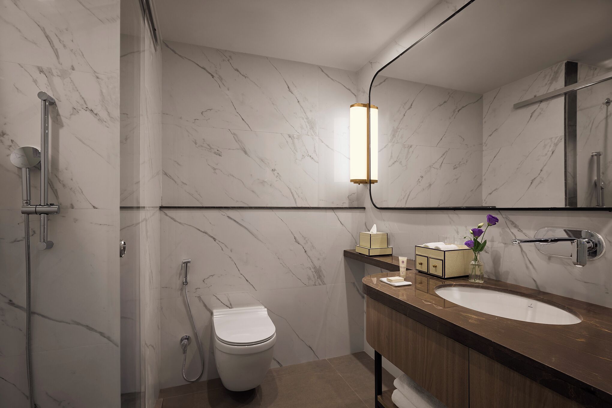 A modern bathroom with a marble wall, a wooden vanity, a white toilet, and a shower area.