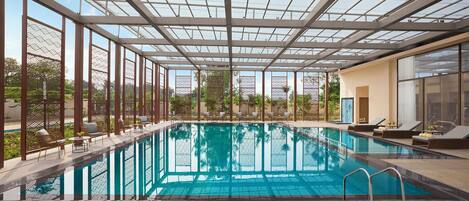 An indoor swimming pool with a glass ceiling, lounge chairs, and a view of the outdoors.
