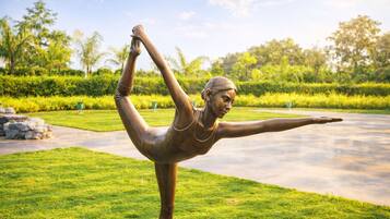 A bronze statue of a person in a yoga pose on a grassy area with a pathway and greenery in the background.