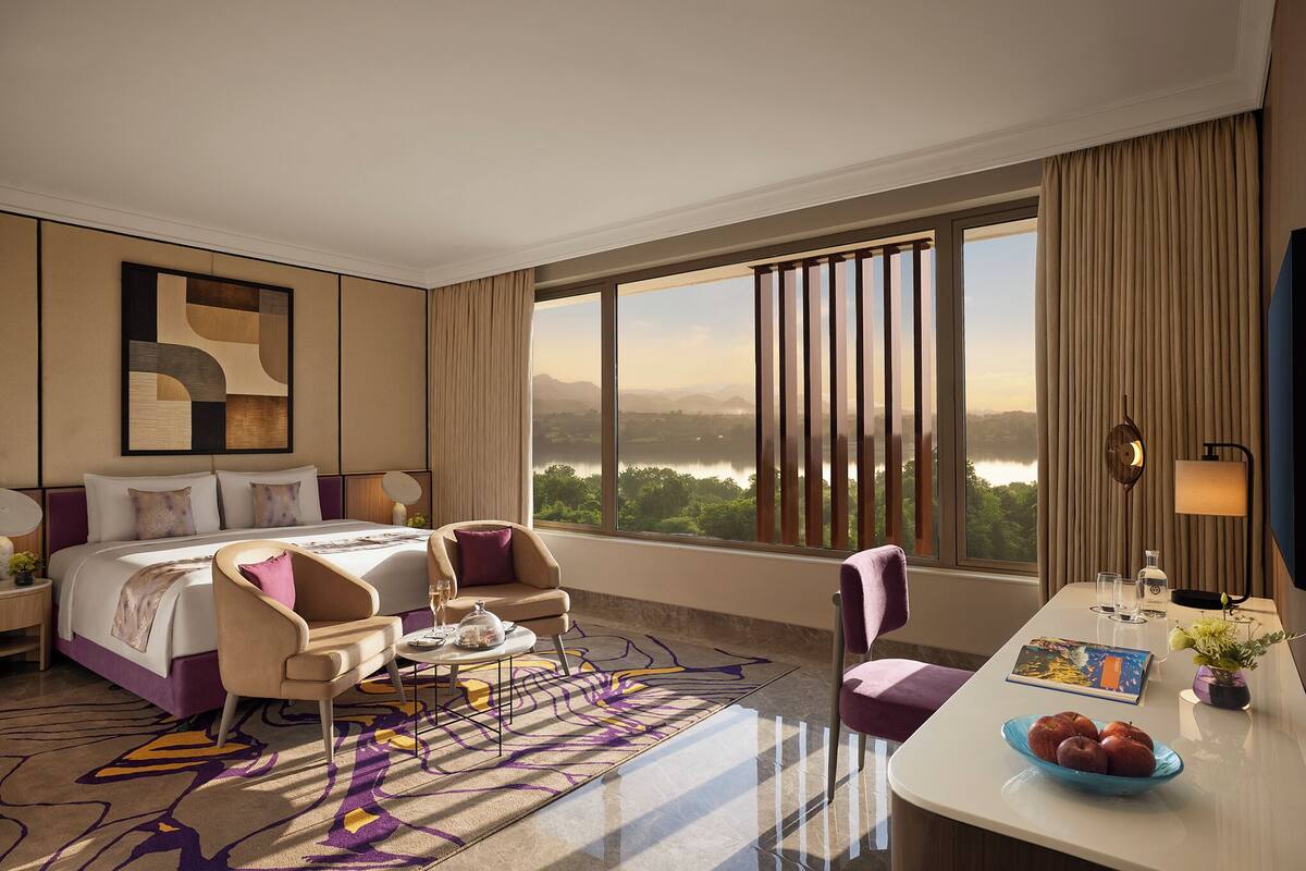 A hotel room with a large bed, two armchairs, a desk with a bowl of apples, and a view of a lake.