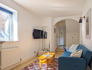 A modern living room with a blue sofa, a wooden coffee table, and a colorful rug. A TV is mounted on the wall, and there is a window with a view of a brick building. - Comfy & Easy 1-Bed Flat in Quiet Maida Vale (London)