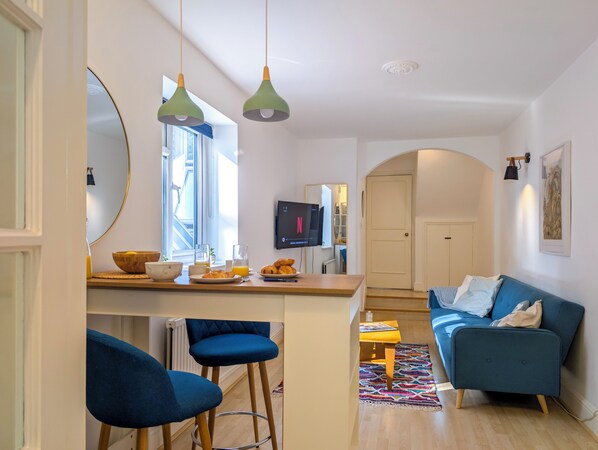 A modern living room with a blue sofa, a small coffee table, and a kitchen area with a dining table and bar stools. - Comfy & Easy 1-Bed Flat in Quiet Maida Vale (London)