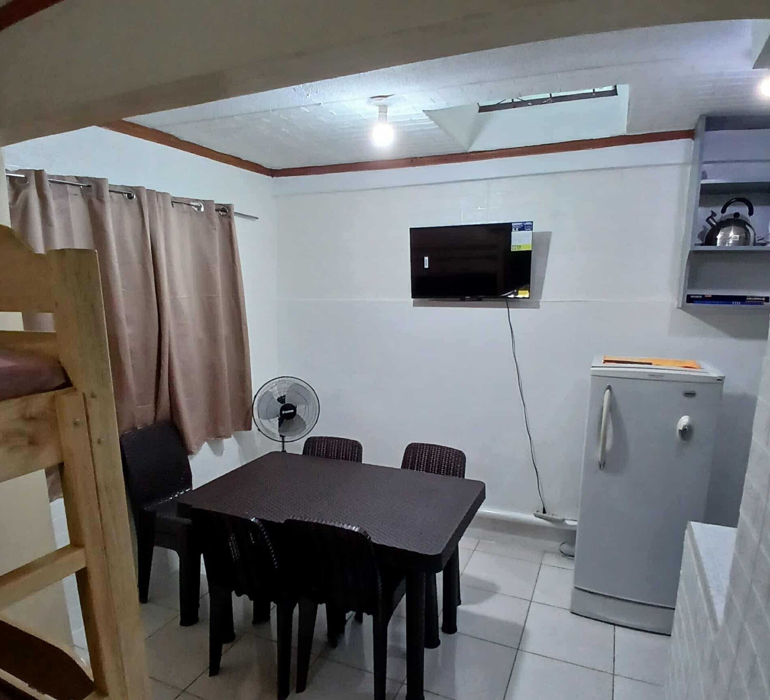 A compact room with a bunk bed, a small table with chairs, a fan, a refrigerator, and a television mounted on the wall.