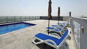 Un espace piscine sur le toit, avec des chaises longues bleues et des parasols, une vue dégagée sur le paysage urbain et une piscine aux carreaux bleus.