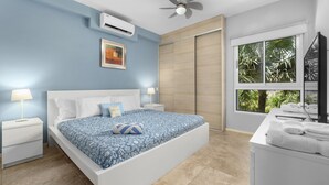 A bedroom with a bed, nightstands, a ceiling fan, an air conditioner, a window with a view of palm trees, and a TV on a stand.