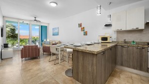 A modern kitchen with a central island, a dining area with a table and chairs, and a large window with a view of palm trees.