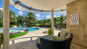 A covered pool area with lounge chairs, a sign with pool rules, and palm trees in the background.