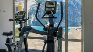 Two stationary bikes with digital displays, positioned in front of a large window overlooking a swimming pool and palm trees.