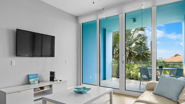 A modern living room with a flat-screen TV, a white coffee table, a sofa, and a sliding glass door leading to an outdoor patio with a table and chairs.