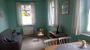 Una habitación con sofá, una mesita, una silla, un radiador, una ventana con cortinas y una planta.
