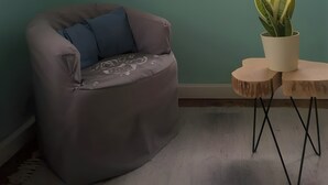 Una habitación con un sillón gris, una mesita de madera con una planta en maceta y una ventana con cortinas.