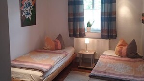Una habitación con dos camas de madera, una mesita con una lámpara, un radiador y una ventana con cortinas.