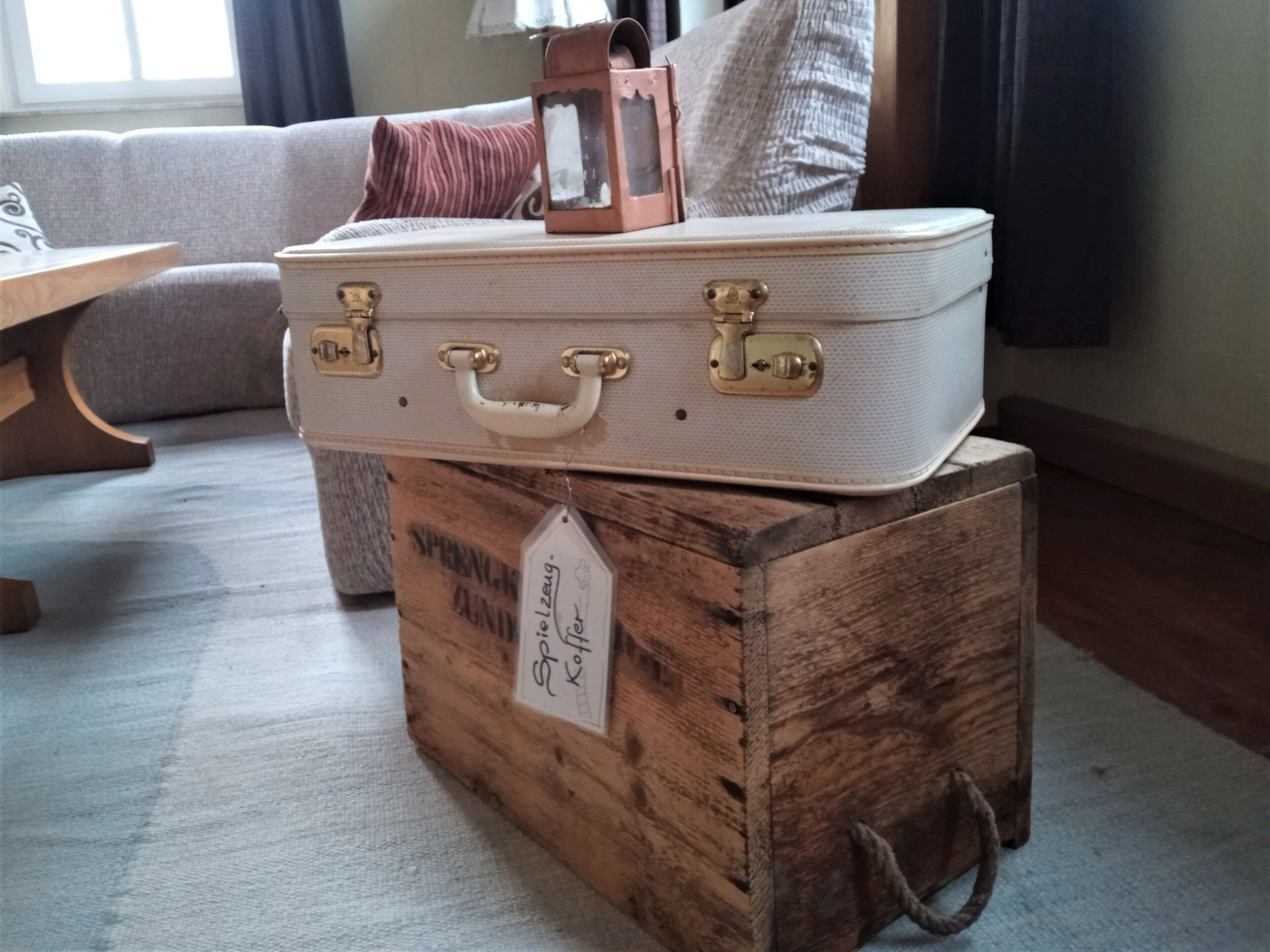 Una maleta antigua sobre un cofre de madera, una etiqueta, un sofá acolchado y una lámpara en una habitación.