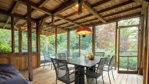 A wooden deck with a glass table and chairs, surrounded by large windows offering a view of trees.